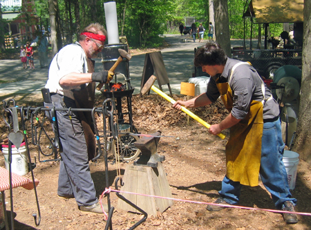 Two  men weild large sledgehammers as they take turns pounding a piece of iron on an outdoor anvil.