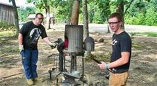 Two men look at the camera while displaying created blacksmith items outdoors.