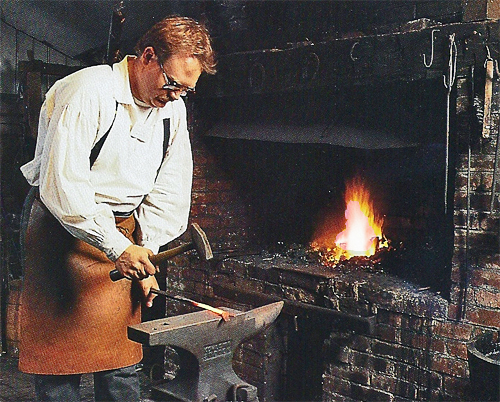 A red hot piece of iron is hammered by a man wearing a blacksmiths apron.  A flaming forge is in the background.