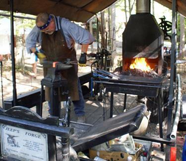 Man in blacksmith apron hammers a red hot piece of iron on an anvil while standing in his portable forge.